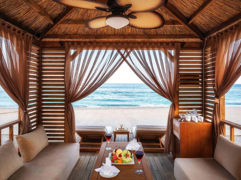 Offene Strandhütte mit Vorhängen, Sofa, Fruchtteller und Blick auf das Meer bei Tageslicht.
