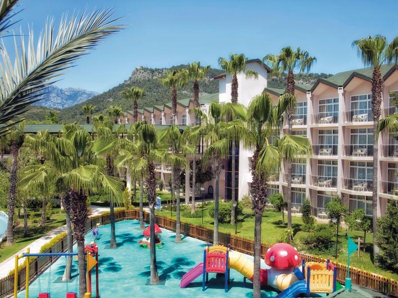 Ein tropischer Garten mit Palmen und einem bunten Kinderspielplatz vor einem Hotelgebäude.