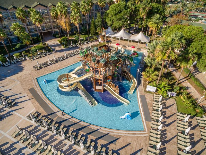 Großer Pool mit Wasserrutsche und vielen Liegestühlen, umgeben von Palmen und grüner Vegetation.