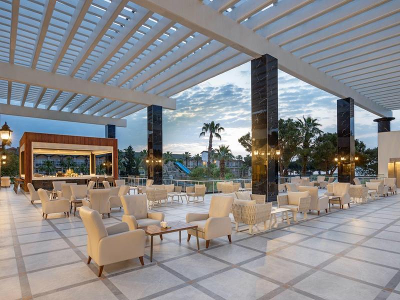 Große, offene Terrasse mit weißen Sesseln, Tischen und moderner Deckenstruktur bei Dämmerung.