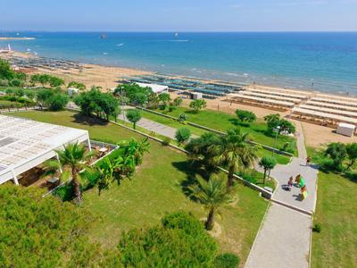 View of a green garden with palm trees, a pathway, and a sandy beach by the sea in the background.
