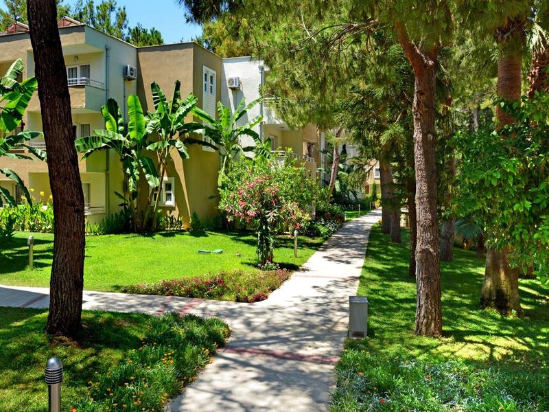Narrow walkway runs through green garden with trees and buildings in hotel area.