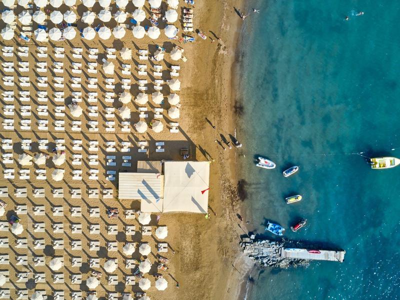 Letecký pohled na pláž s řadami slunečníků a lehátek vedle čisté modré vody a lodí.