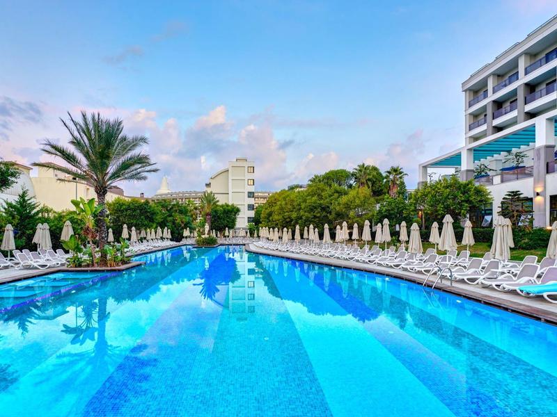 Velký venkovní bazén s lehátky a palmami před budovami hotelu za jasné oblohy.