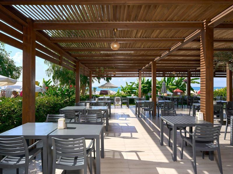 Venku jídelní prostor s dřevěnou střechou, šedými stoly a židlemi, obklopený rostlinami.