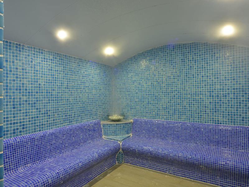 Parní místnost s modrými mozaikovými dlaždicemi a vestavěnými rohovými lavicemi.