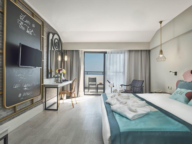 Světý hotelový pokoj s velkou postelí, posezením a balkonem s výhledem na moře.