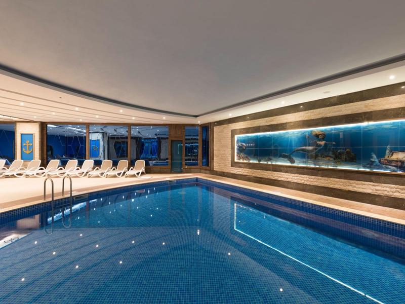 Piscina cubierta moderna con tumbonas y gran muro de vidrio en área de hotel lujoso.