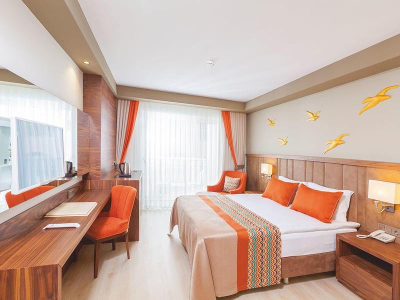 Helles Hotelzimmer mit Doppelbett, orangefarbenen Akzenten und Schreibtisch am Fenster.