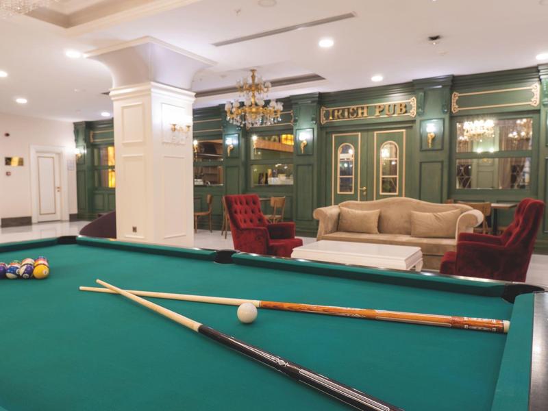 Ein grüner Billardtisch mit Kugeln und Queues in einem Raum mit Sessel, Sofa und Bücherregal.