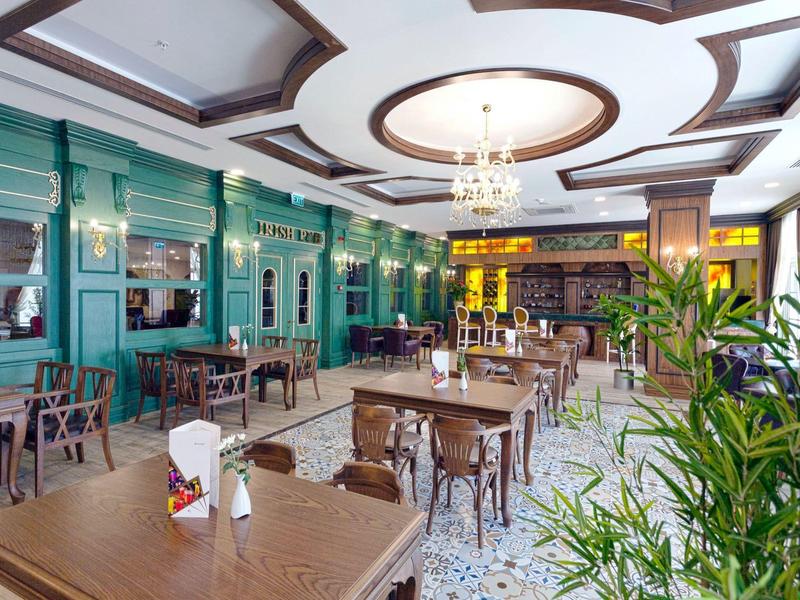 Eleganter Gastraum mit grünen Wänden, Holztischen, Kronleuchtern und gepflanzter Dekoration.