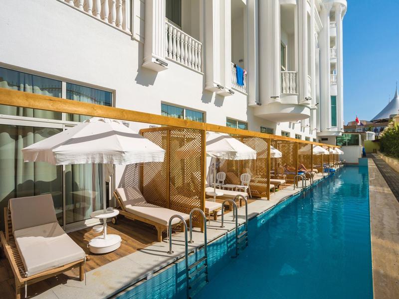 Schwimmbad mit Liegestühlen und Sonnenschirmen an einem weißen Gebäude bei klarem Himmel.
