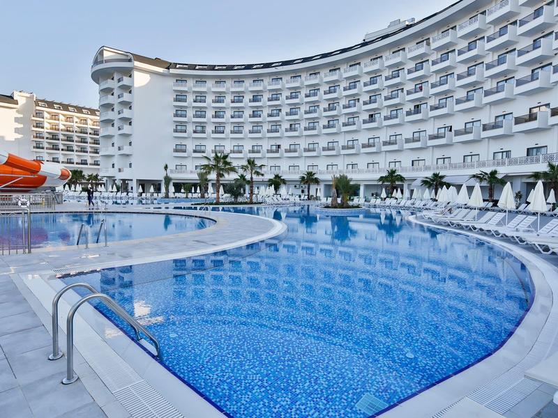 Grand bâtiment d'hôtel courbé avec piscine extérieure et toboggan sous un ciel bleu clair.