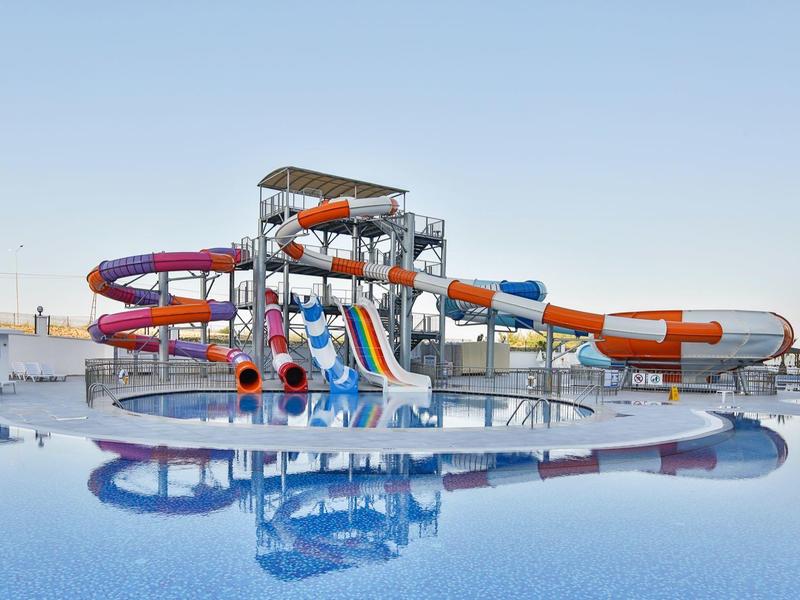 Parc aquatique avec toboggans tubulaires colorés et piscine calme dans la zone hôtelière sous ciel dégagé.