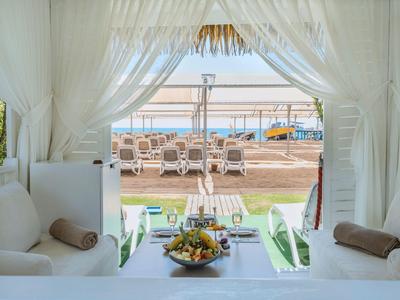 Vista desde una habitación blanca y acogedora con cortinas hacia una playa con tumbonas y el mar.