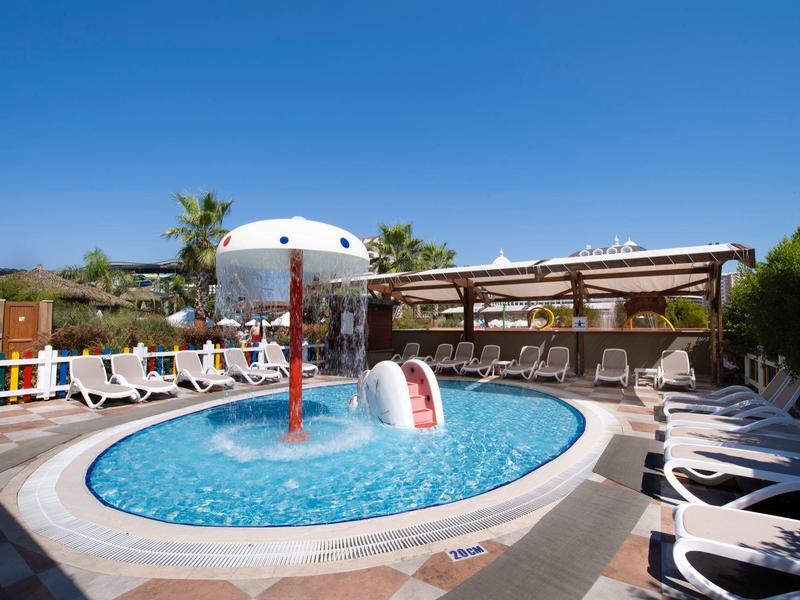 Kruhový dětský bazén s tobogánem a slunečníky, obklopený lehátky pod modrou oblohou.