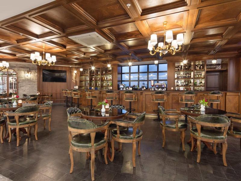 Elegant hotel bar room with wooden ceiling, chandeliers, and round tables with green chairs.