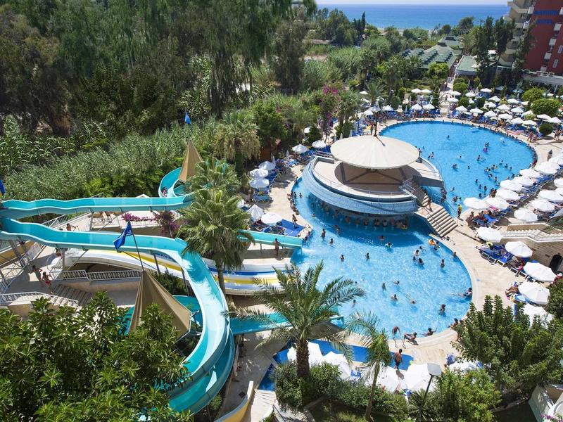 Large outdoor pool with water slides and umbrellas, surrounded by trees and hotel buildings.