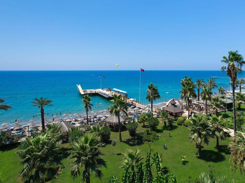 View of a hotel garden with palm trees and beach with a pier by the sea.
