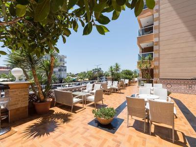 Sunny outdoor terrace with tables, chairs, plants, and a view of nearby buildings and parked cars.