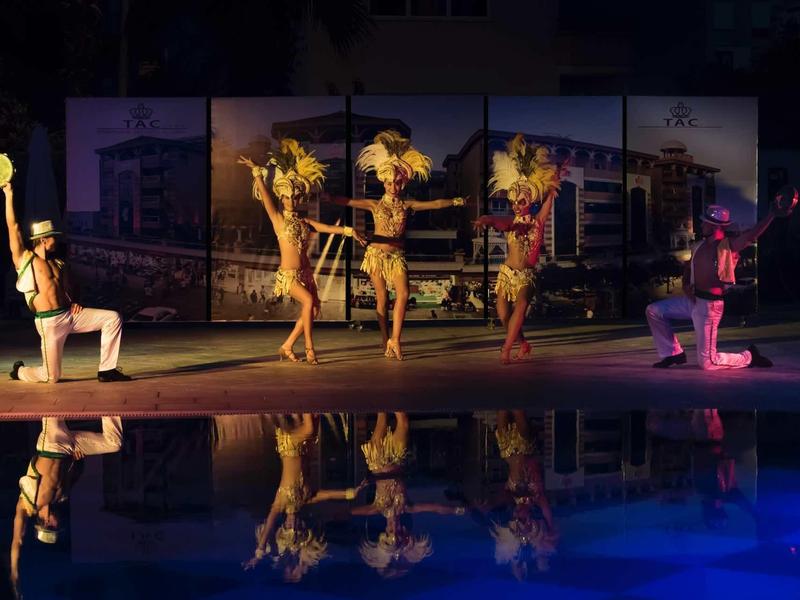 Performers dance and pose near a pool at night with colorful lighting and reflections in the water.