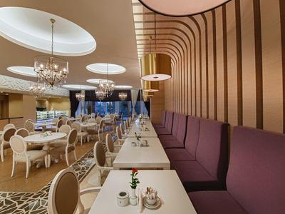 Moderner Speiseraum mit weißen Tischen, lila Sitzbänken, Kronleuchtern und rundem Deckenlicht.