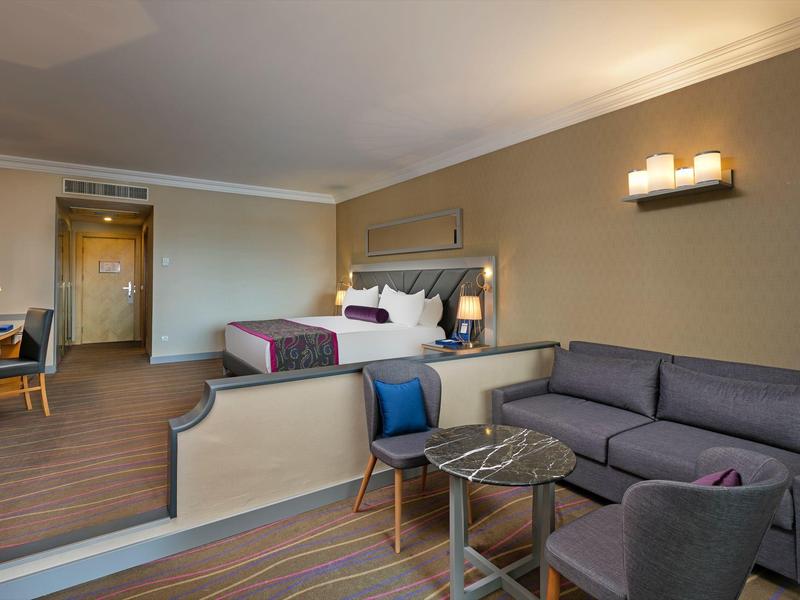 Modernes Hotelzimmer mit Doppelbett, grauer Couch, Sessel, Tisch und Schreibtisch, warm beleuchtet.