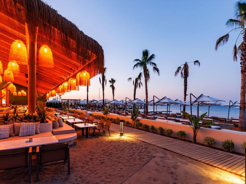 Strandrestaurant bei Sonnenuntergang mit leuchtenden Laternen, Palmengarten und Meerblick.