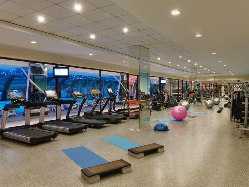 Großer Fitnessraum mit Laufbändern, Gewichten, Gymnastikbällen und Steppern, gut beleuchtet.