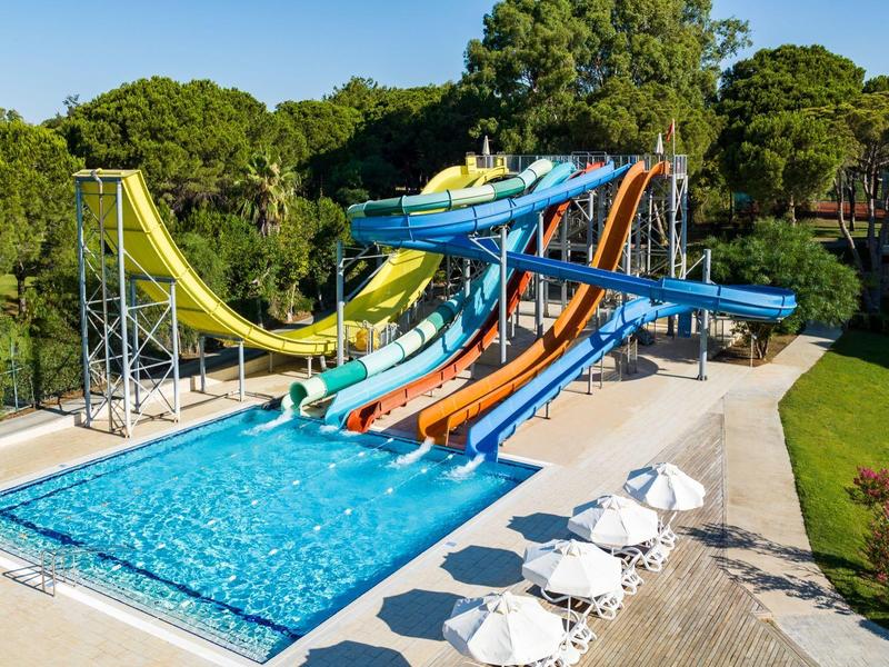 Mehrere bunte Wasserrutschen führen in ein Schwimmbecken, umgeben von grünen Bäumen und weißen Sonnenschirmen.