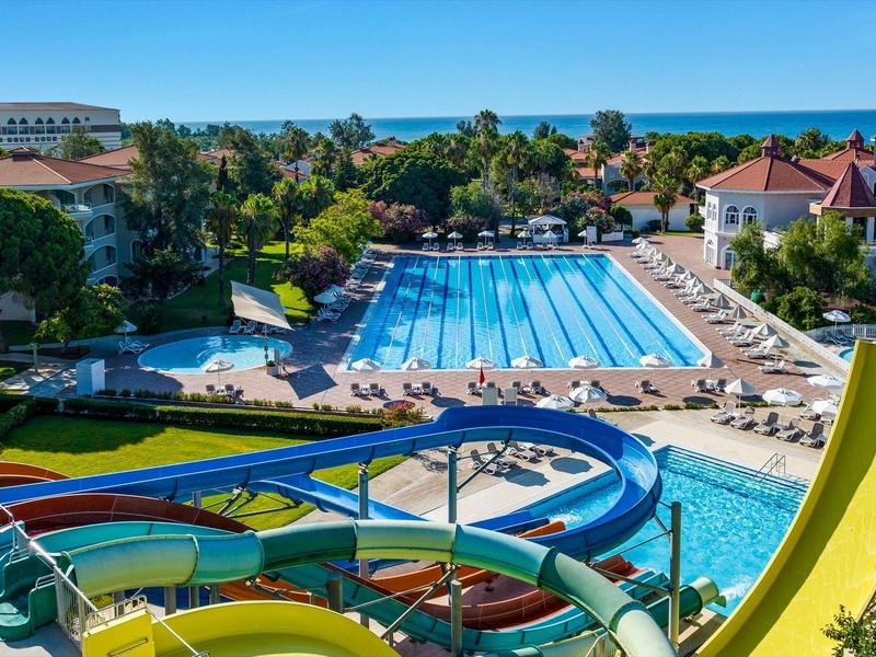 Großer Außenpool mit mehreren Wasserrutschen, Sonnenterrasse und mediterranem Hotel im Hintergrund.