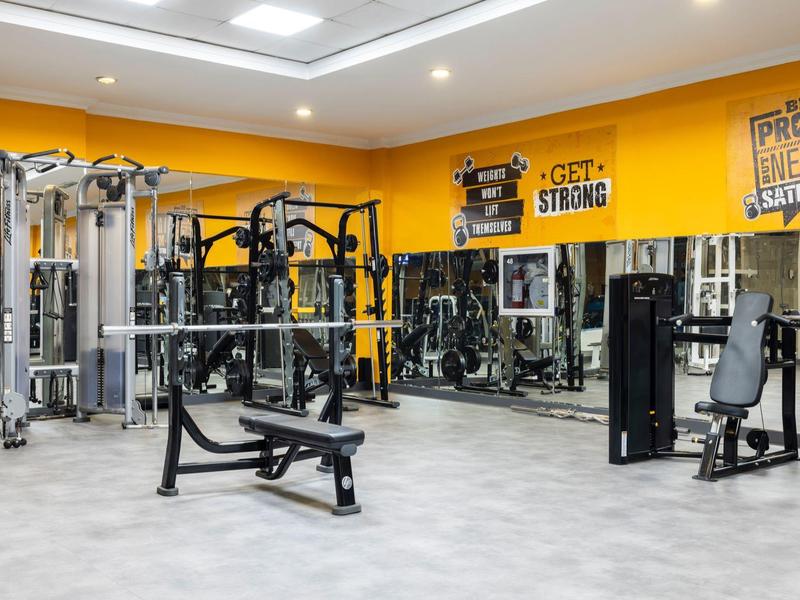 Moderne Fitnesshalle mit gelben Wänden, vielfältigen Maschinen und Spiegelflächen.