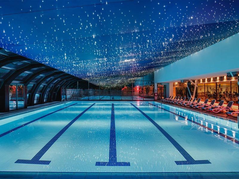 Schwimmbad mit beleuchtetem Sternenhimmel, blauer Wasserfläche und Sitzbereich rechts.