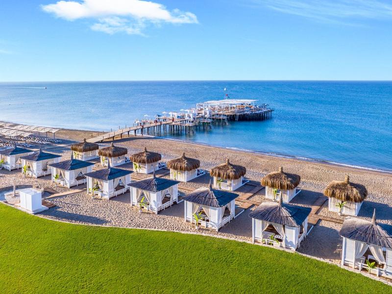 Strand mit Reihen von weißen Strandhäuschen, Liegen, grünem Rasen und Holzpier im blauen Meer.