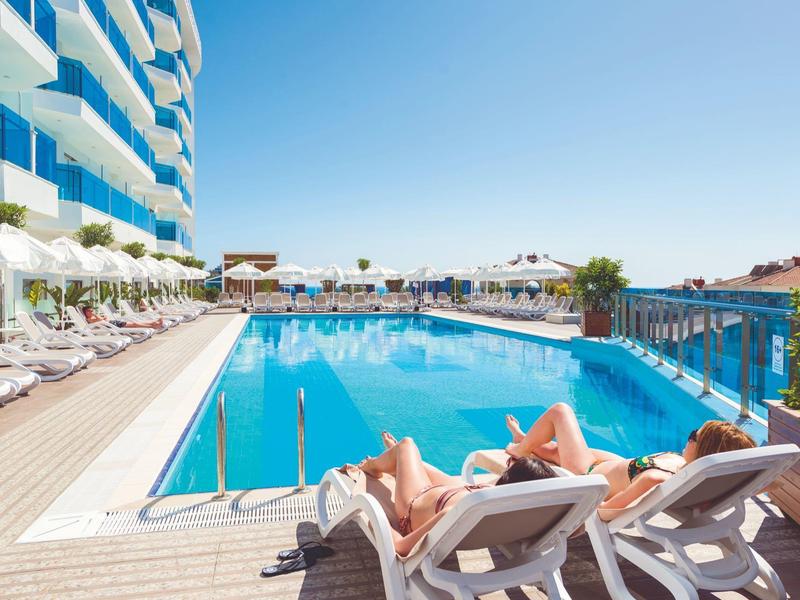 Hotel pool with sun loungers and guests, modern building and clear blue sky.