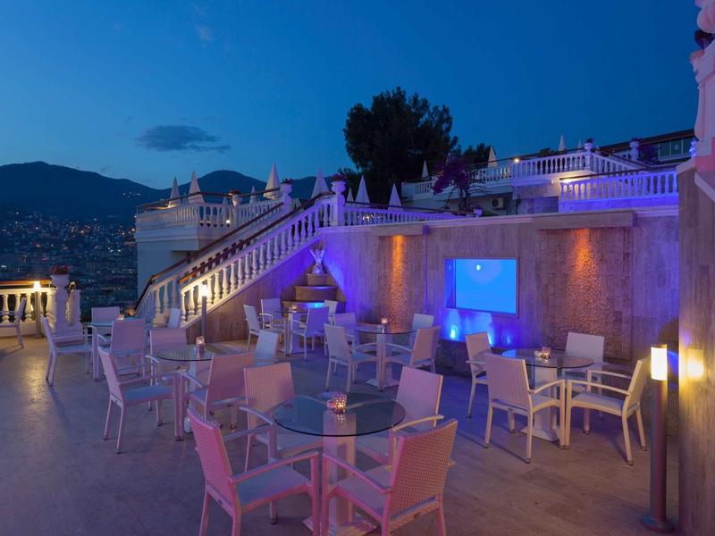 Venkovní terasa hotelu s stolky a židlemi, večerním osvětlením a výhledem na hory.