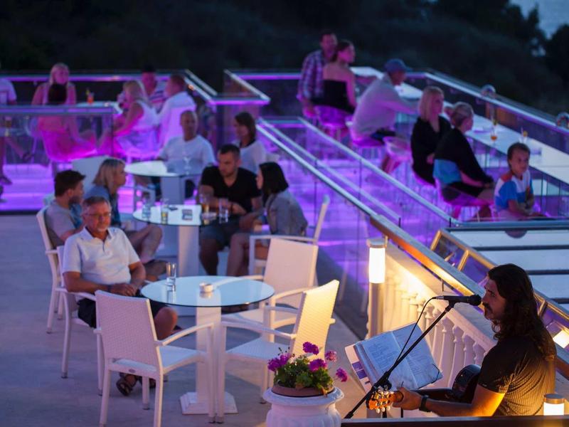 Večerní lounge se sezením, skleněnou podlahou a hudebníkem na pozadí kopců.