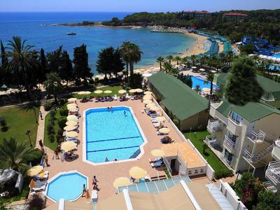 Blick auf einen Hotelpool mit Sonnenschirmen und Garten, dahinter Strand und blauer Meereshorizont.