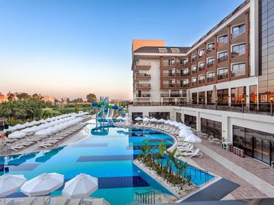 Moderner Hotelpool mit blauen Wasserflächen, weißen Sonnenschirmen und angrenzendem Gebäude bei Sonnenuntergang.