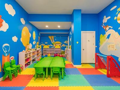 Bunter, heller Kinderspielraum mit Wandbildern, bunter Matte, Tischen, Stühlen und Regalen voller Spielzeug.