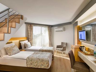 Moderner Hotelzimmer mit zwei Einzelbetten, Holztreppe, Schreibtisch und großen Fenstern mit Vorhängen.