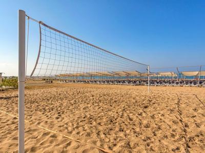 Leeres Volleyballnetz auf sonnigem Sandstrand mit blauem Himmel und Sonnenschirmen im Hintergrund.