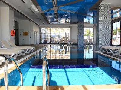 Helles Hallenbad mit blauen Wasser, weißen Liegen und großen Fenstern für viel Tageslicht.
