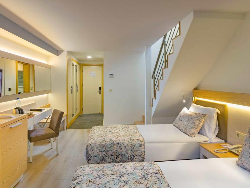 Modern hotel room with two single beds, desk, chair, and staircase in the background.