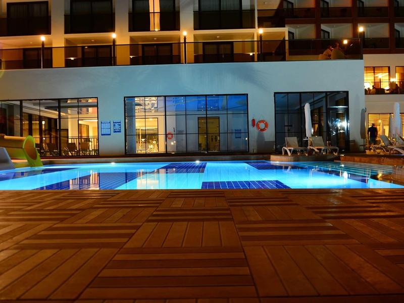 Illuminated hotel pool at night with wooden deck and modern facade in background