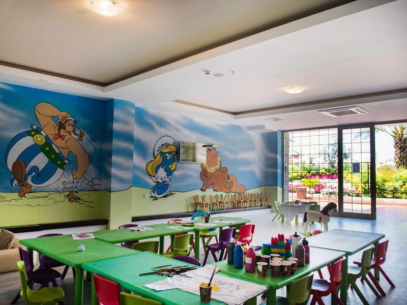 Kinderzimmer mit bunten Tischen, Stühlen und Wandbildern von Comicfiguren und Landschaft.