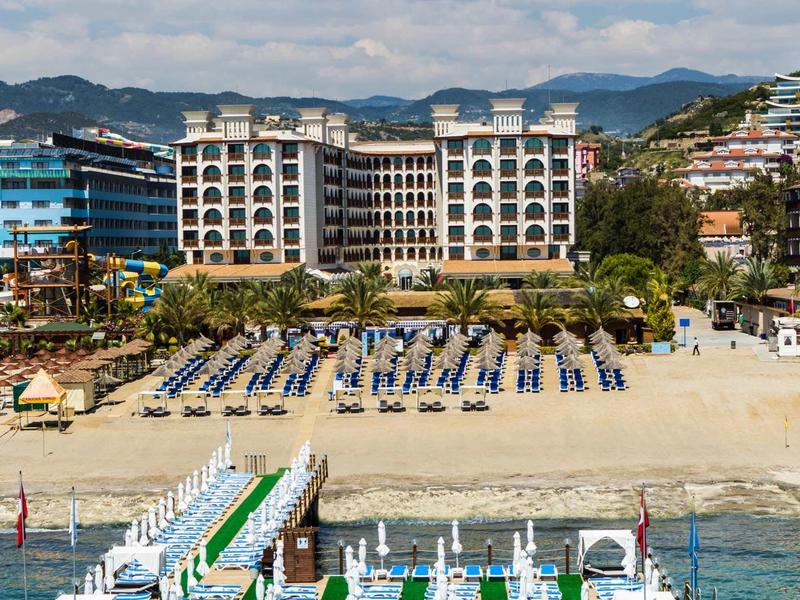 Hotel am Sandstrand mit Liegen, Steg und klarem Meer, im Hintergrund Berge und bewölkter Himmel.