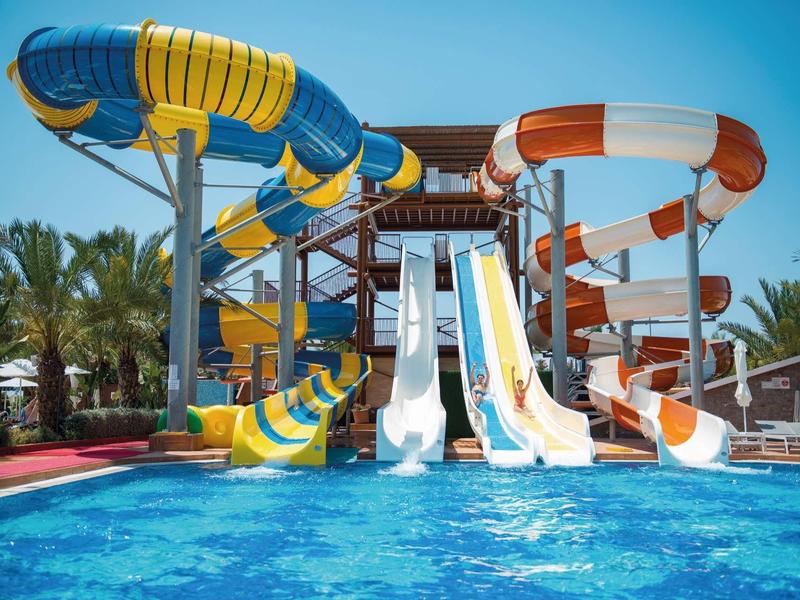 Bunte Wasserrutschen führen von einer Plattform in ein klares blaues Schwimmbecken unter blauem Himmel.