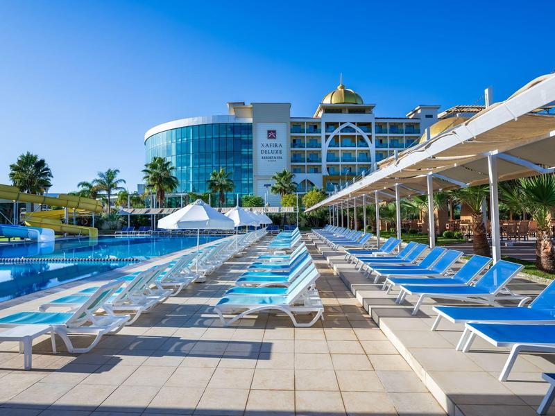 Velká oblast s bazénem a modrými lehátky, hotelová budova v pozadí za jasné oblohy.