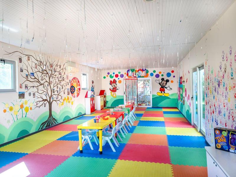 Barevná herna s stoly, židlemi a veselými malbami na stěnách pro děti.